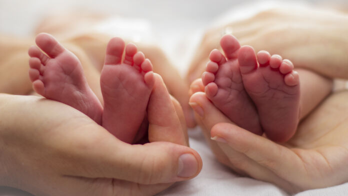 Mother and father's hands cradling twin babies' feet on a pale background.